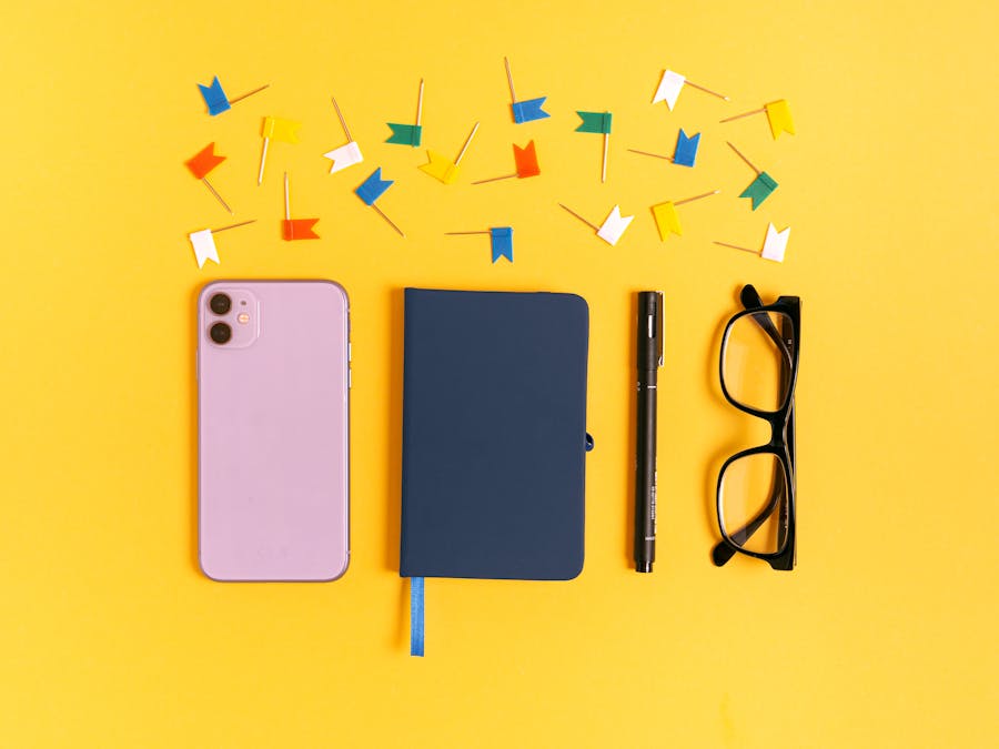 A vibrant flat lay of a smartphone, notebook, pen, eyeglasses, and colorful pins on a yellow background.