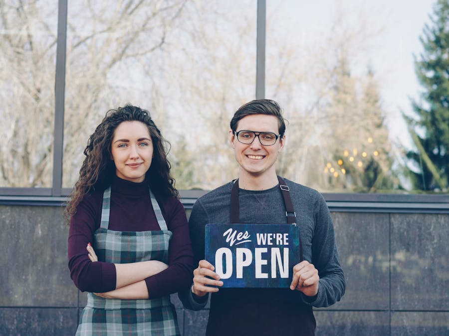 Two business owners standing outside, holding a 'Yes We're Open' sign, welcoming customers.