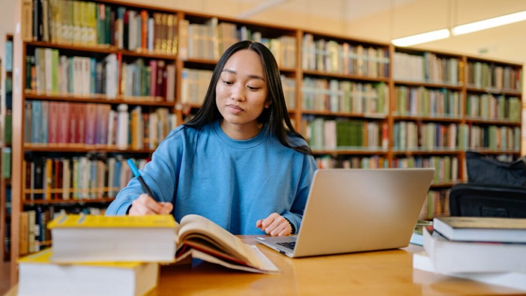 Young woman studying in a library with books and laptop, focusing on education.