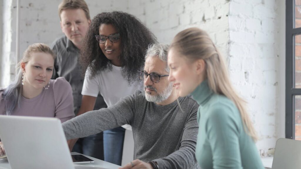 Group of diverse colleagues sitting and standing near table with laptop while discussing project and working together during business meeting