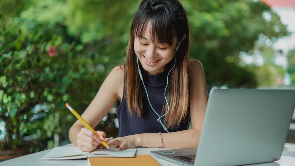 Positive young Asian female student with earphones writing in copybook while doing homework at table with laptop in street cafeteria