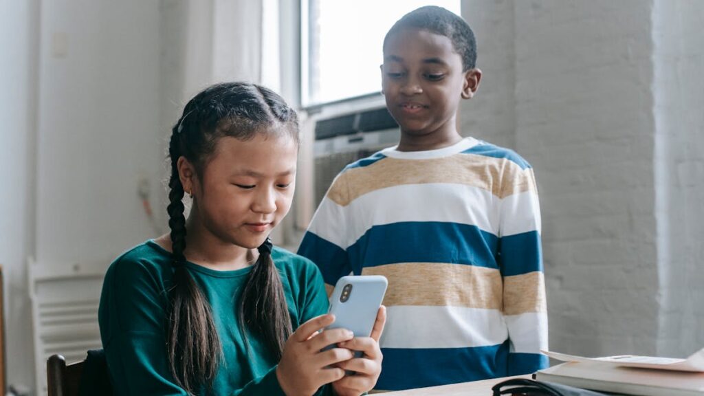 Happy little African American boy standing near Asian classmate girl using smartphone sitting at desk during break