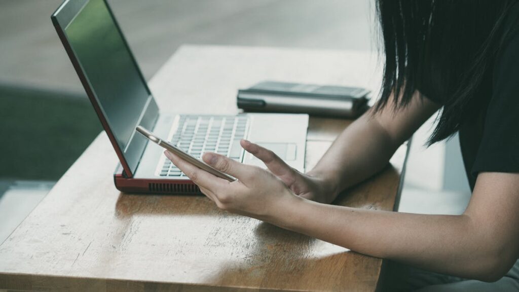 A young woman multitasking with her smartphone and laptop at a table indoors.