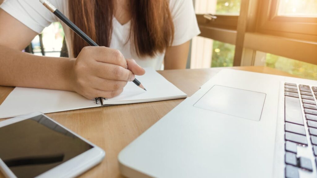 A young woman writing notes at a table with laptop and smartphone.