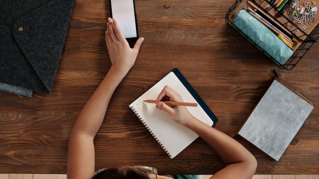 Overhead view of a child writing in a notebook while using a smartphone at a wooden desk.