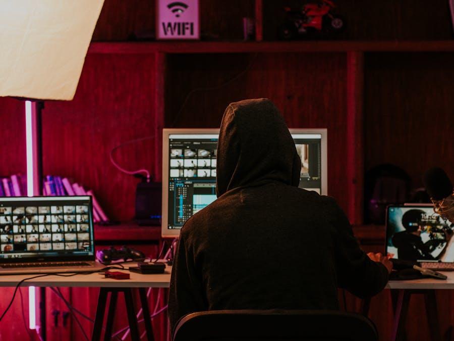 Person in a hoodie editing videos on multiple screens in a dimly lit space with neon lighting.