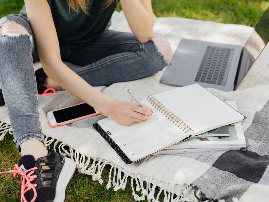 Crop faceless female student in casual clothes taking notes in planner while sitting on blanket with smartphone and laptop on green park grass on sunny day