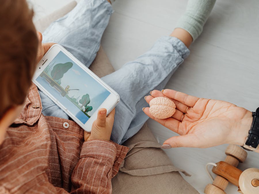 Young child engrossed in a smartphone video while holding a brain model indoors.