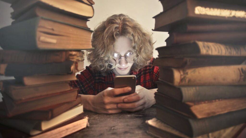 Focused male student in casual wear and glasses browsing smartphone while sitting at wooden table with stacked old books