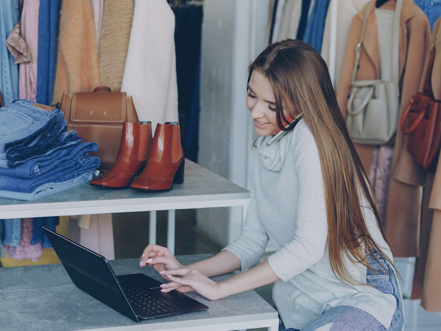 Young woman managing a fashion boutique, multitasking with phone and laptop, surrounded by clothing.