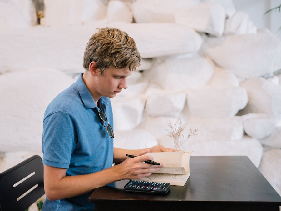 A young adult male in a blue polo shirt studies at a table indoors, using a book and smartphone.