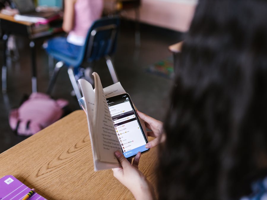 A student in a classroom multitasks by reading a book while using a smartphone, emphasizing modern educational challenges.