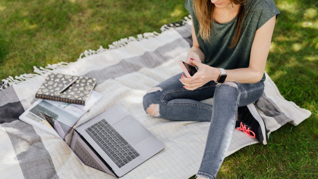 From above of young crop female student in casual clothes surfing social media on smartphone while sitting with laptop and notebooks on blanket placed on green grass in sunny park