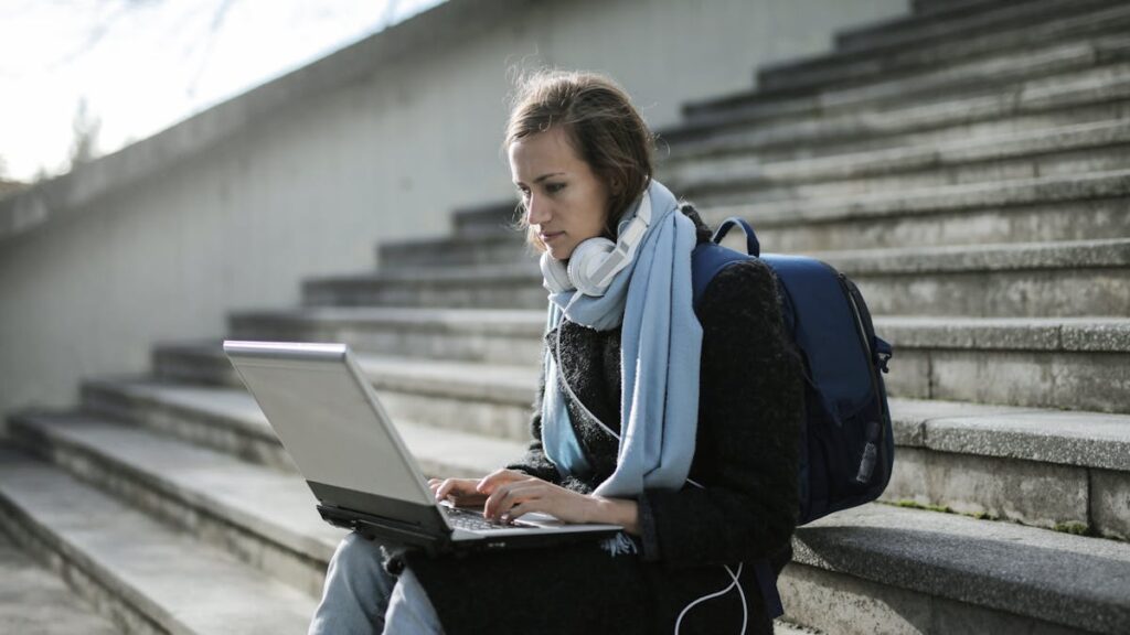 Woman using laptop on outdoor stairs, studying online with headphones and backpack.