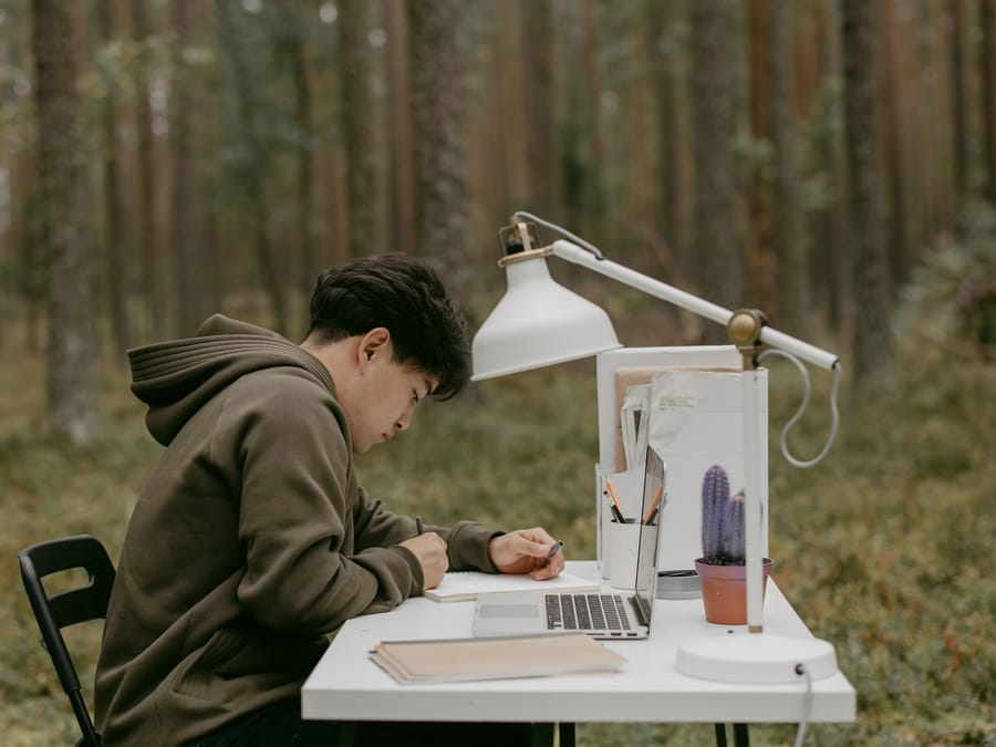 A person studying outside using a laptop and notebook in a natural setting. Ideal for education or remote work concepts.