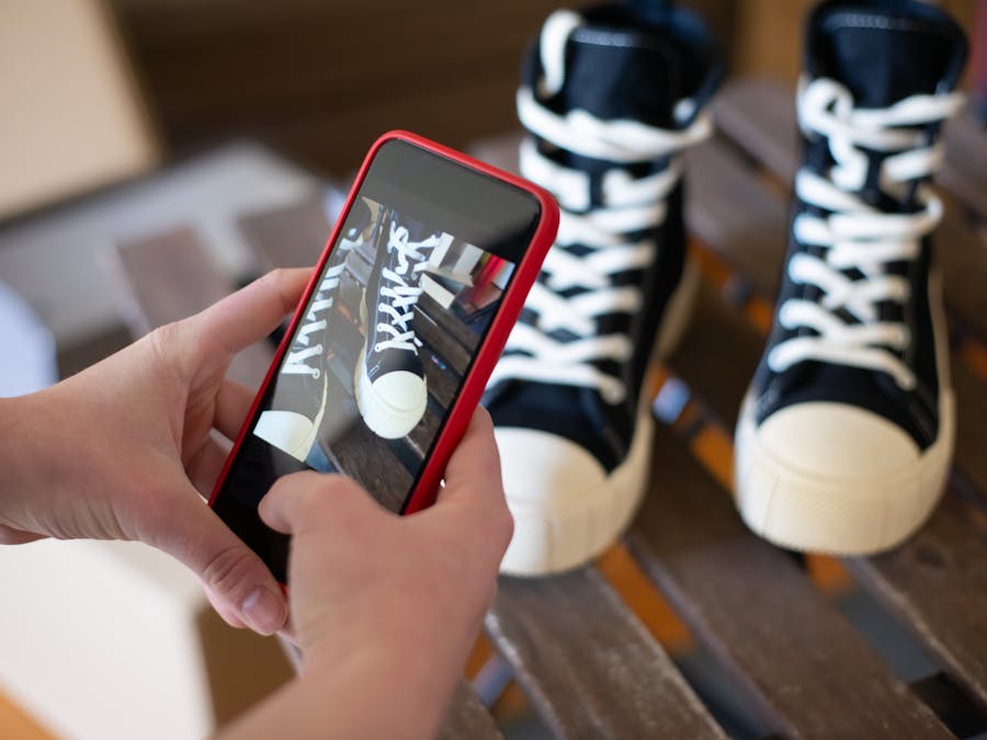 Hands using a smartphone to photograph black sneakers on a wooden surface.