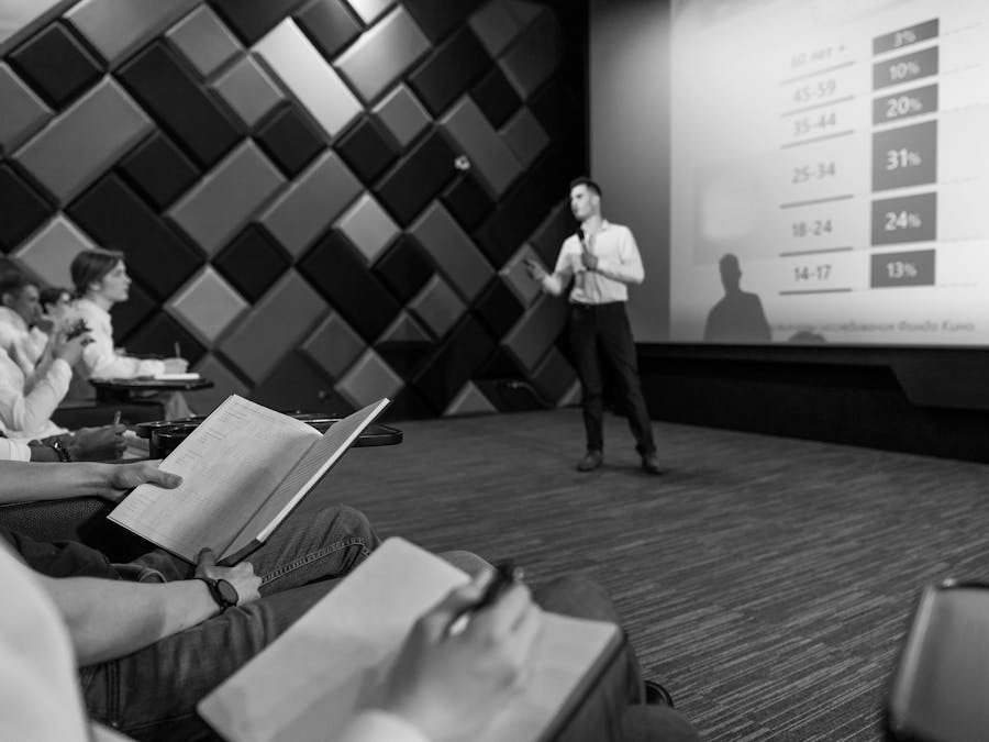 Monochrome image of a business presentation with an audience taking notes.