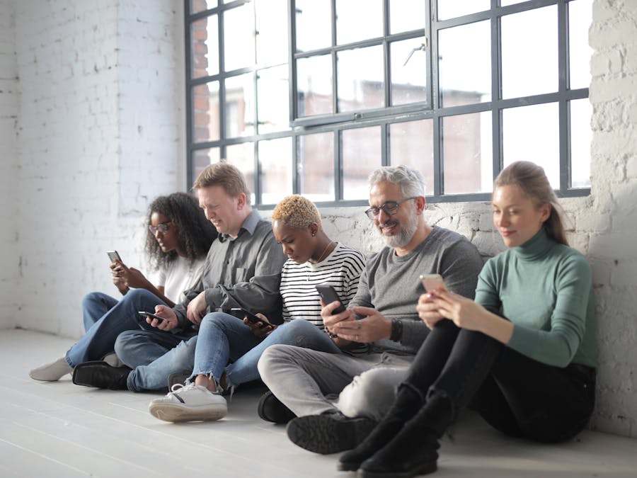 Smiling coworkers of different ages in casual outfit browsing internet on cellphones while sitting with crossed legs near brick wall and fenced window during daytime