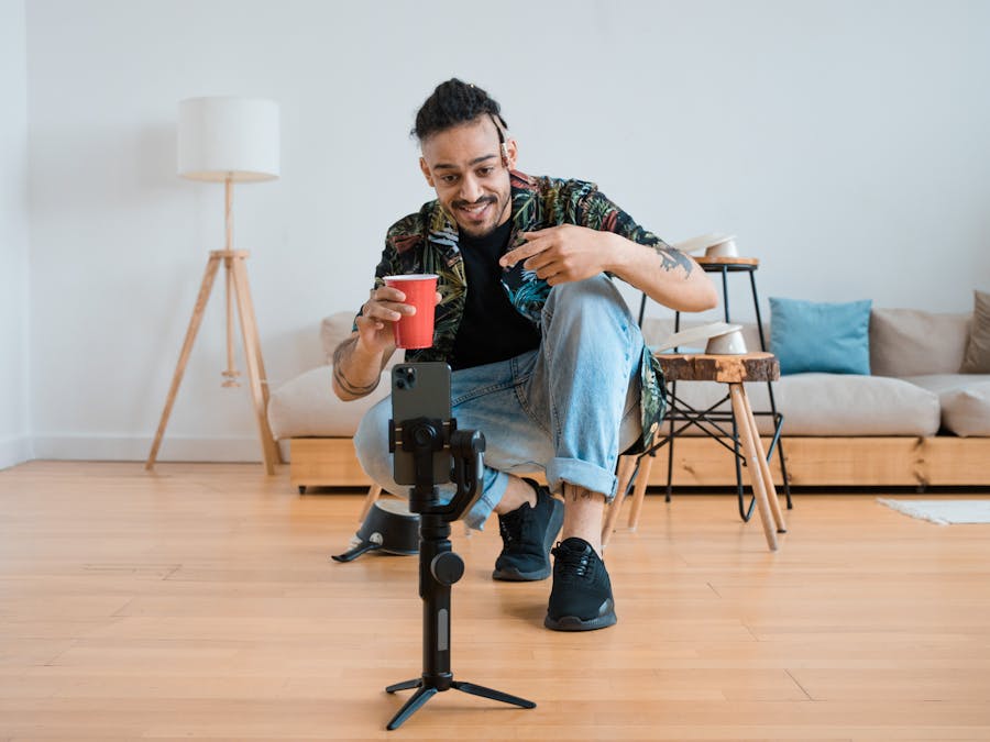 Man filming vlogs in a living room setting, holding a red cup and gesturing at smartphone on tripod.