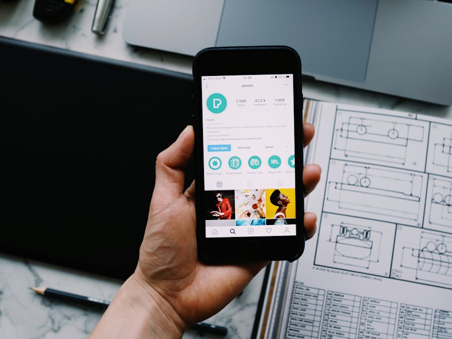 Hand holding smartphone displaying social media profile with tech gadgets on desk.
