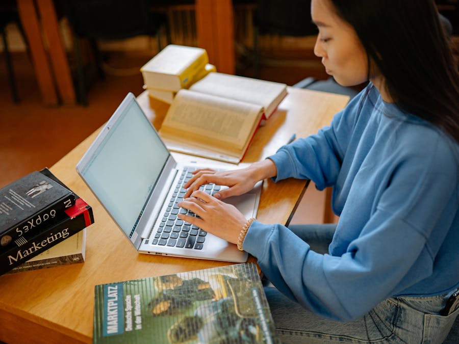 Young woman typing on a laptop surrounded by textbooks in a library setting.