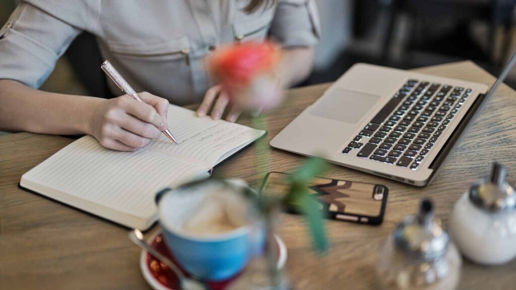 Person writing content ideas in a notebook beside a laptop