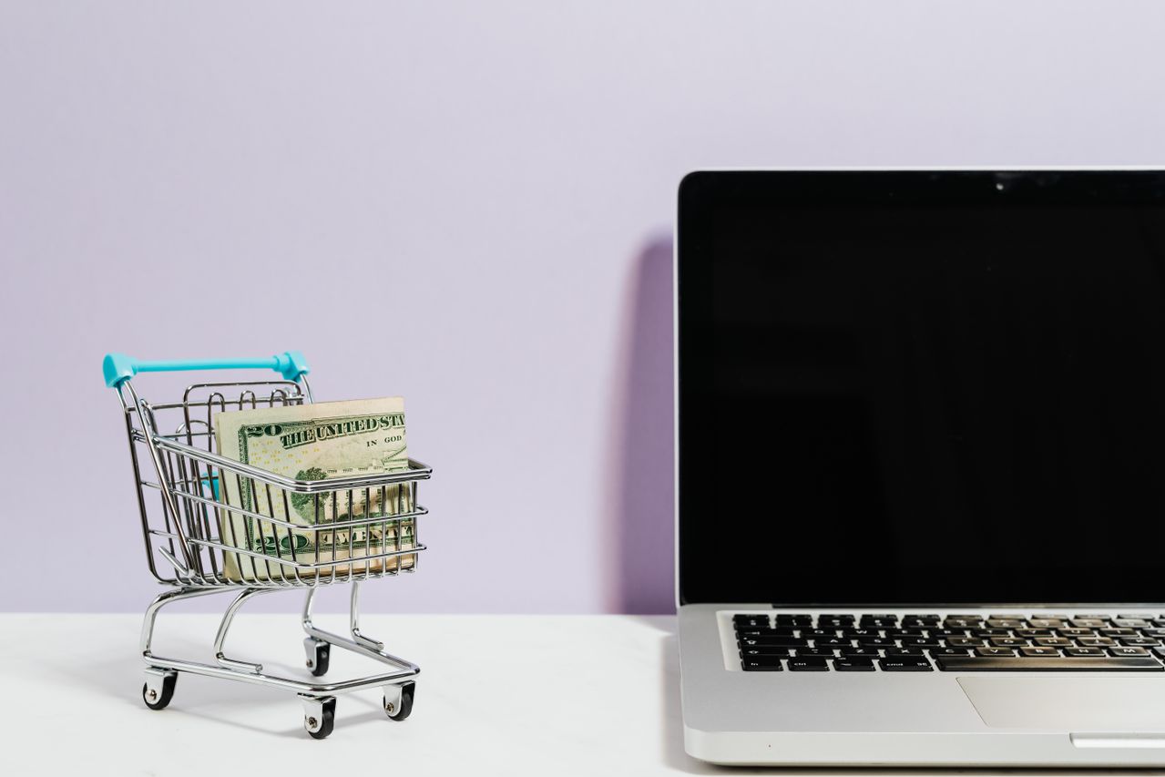 Miniature shopping cart holding cash beside an open laptop