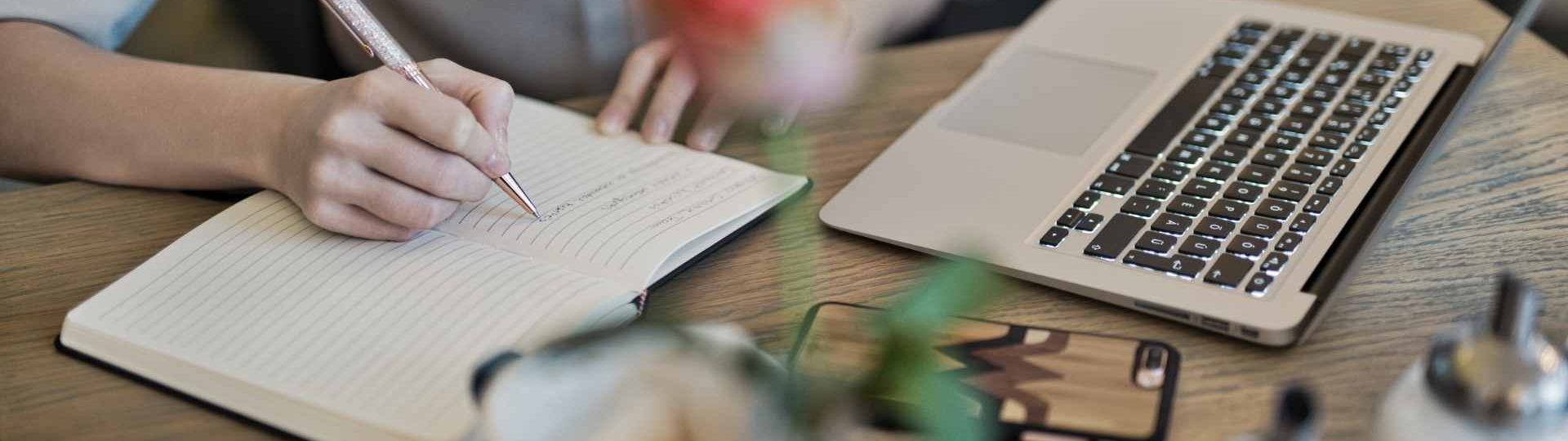 Person writing content ideas in a notebook beside a laptop