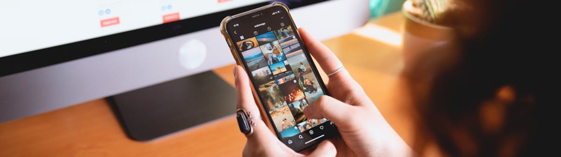 A person browsing images on a smartphone while sitting at a desk with a desktop monitor.