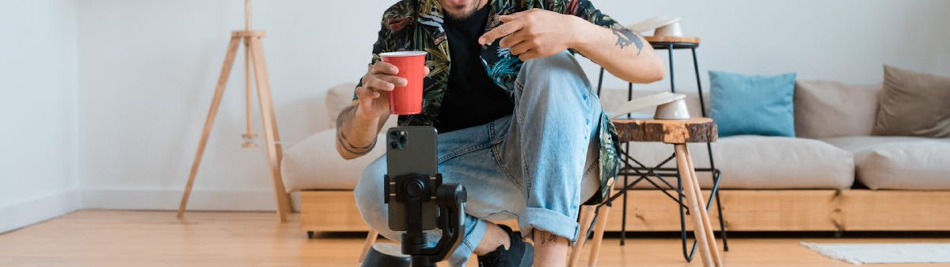 Man filming vlogs in a living room setting, holding a red cup and gesturing at smartphone on tripod.