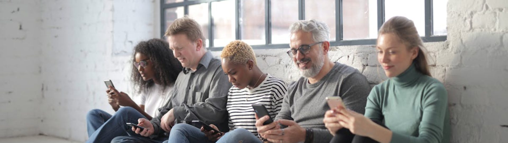 Smiling coworkers of different ages in casual outfit browsing internet on cellphones while sitting with crossed legs near brick wall and fenced window during daytime