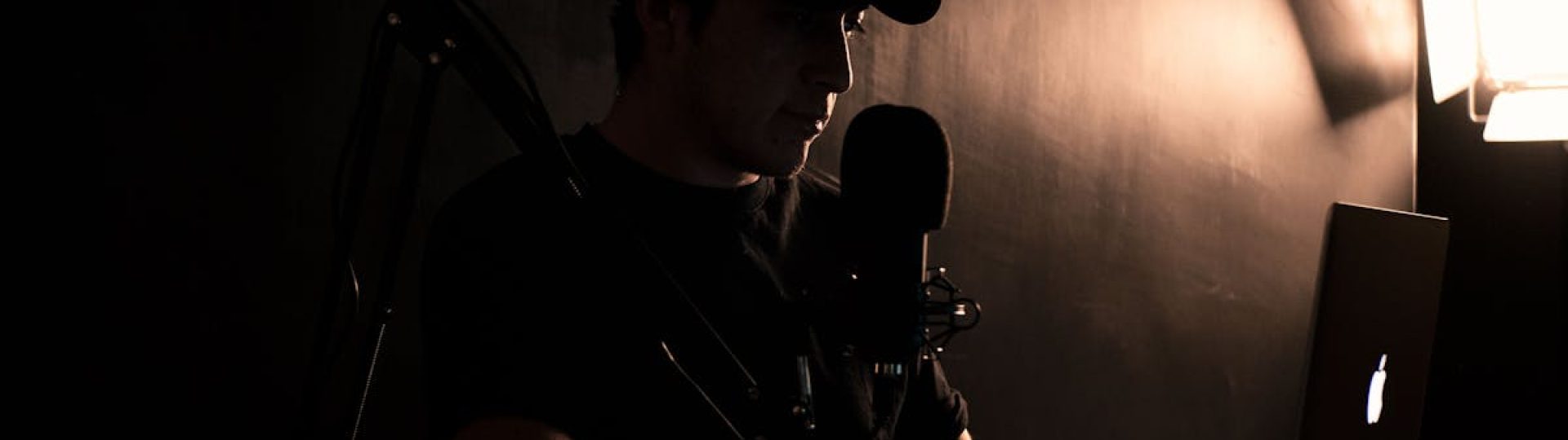 A man wearing a cap works on a laptop with a microphone setup in a dimly lit room.