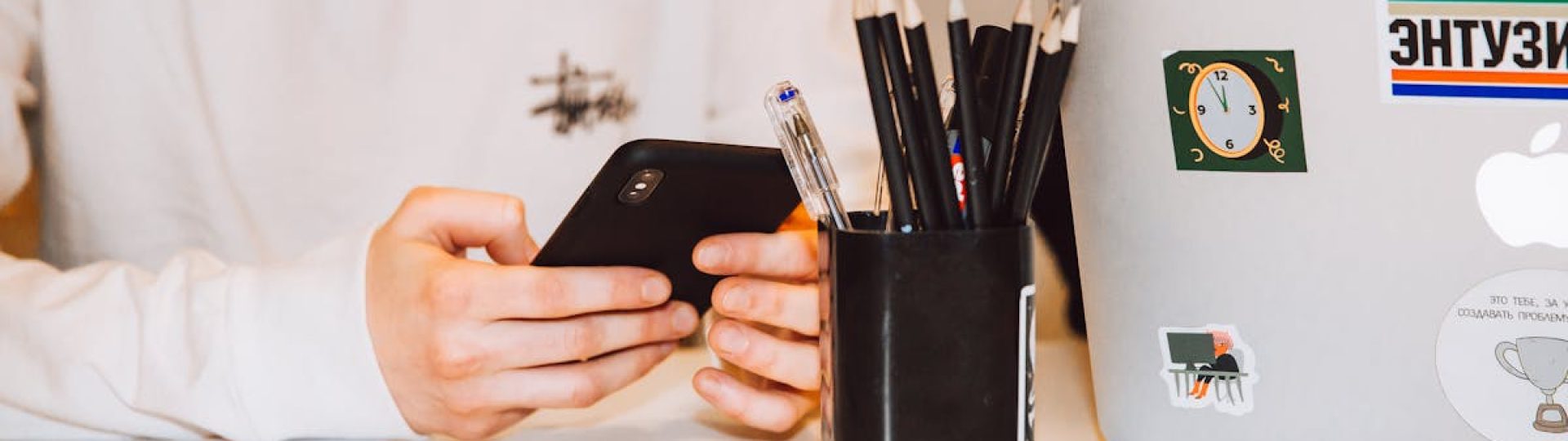 Young adult at a desk texting on smartphone, next to a laptop and stationery holder.