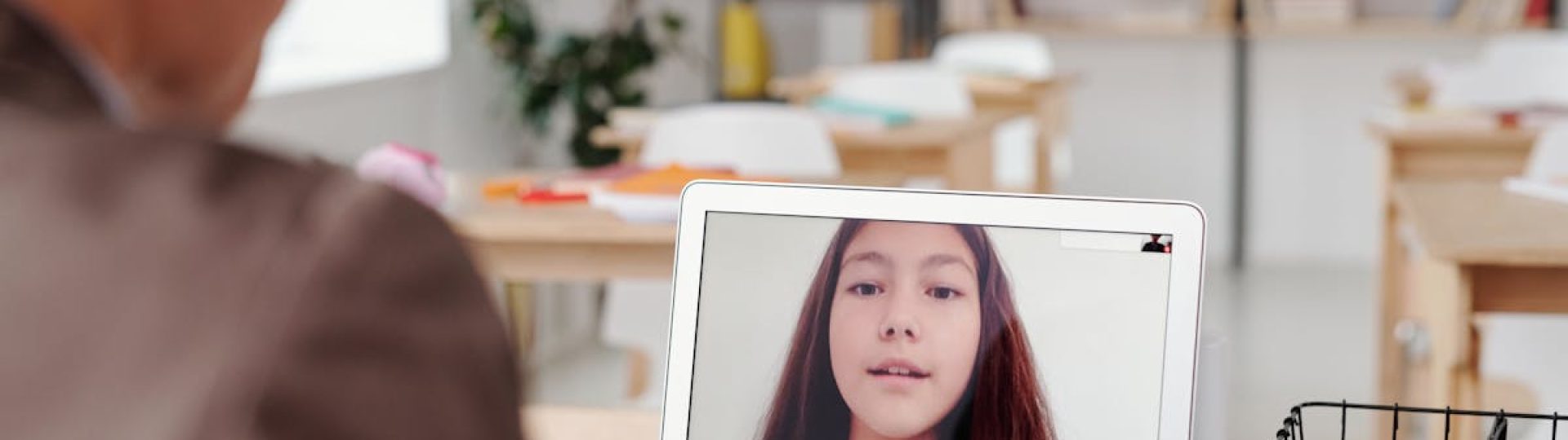Teacher using laptop for virtual lesson with female student on screen in classroom setting.