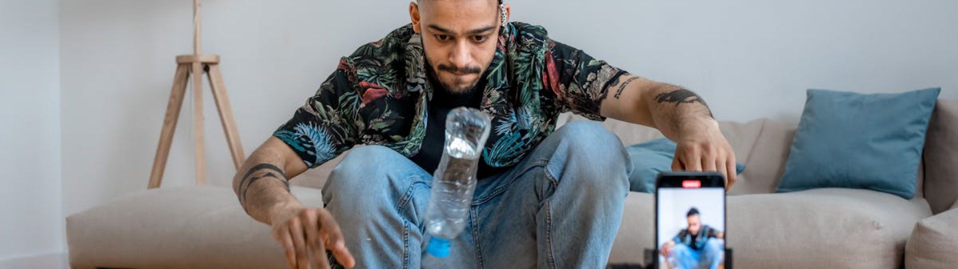 A young man focuses on a bottle flip challenge while filming content at home, capturing it on a smartphone.