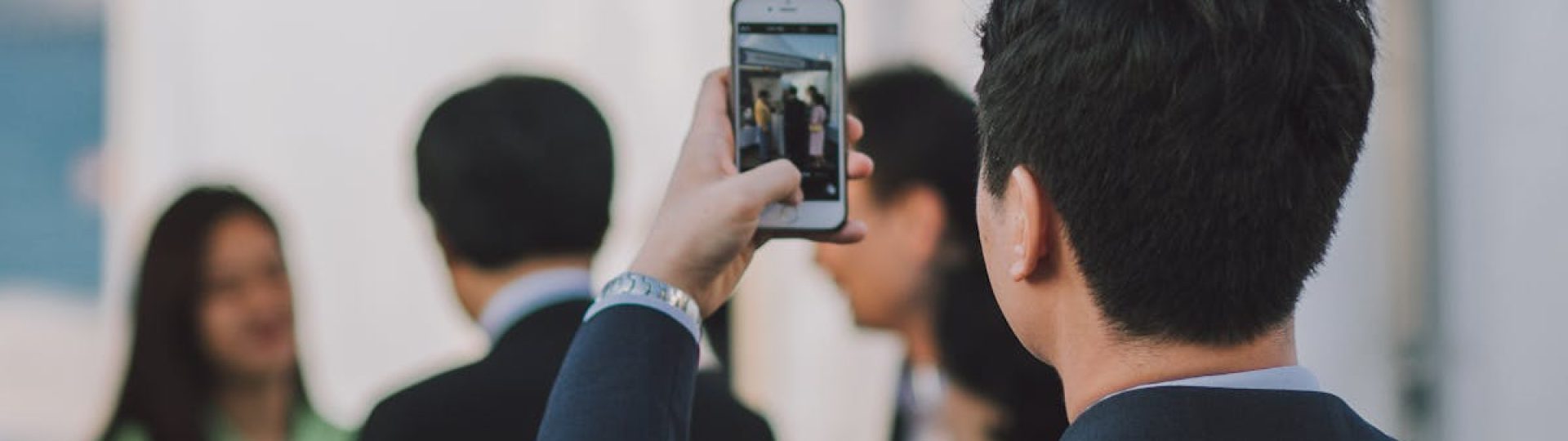 Man in suit photographing colleagues with smartphone during business event outdoors.