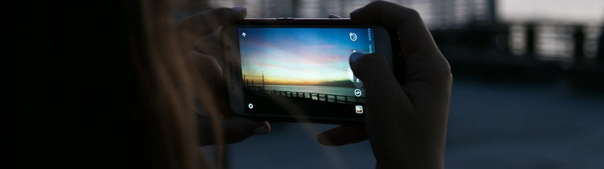 A person photographs a vibrant sunset with a smartphone by the waterfront. Outdoor scene.