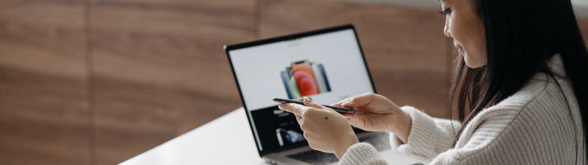 Young Asian woman setting up a new smartphone while working on a laptop in a modern indoor setting.