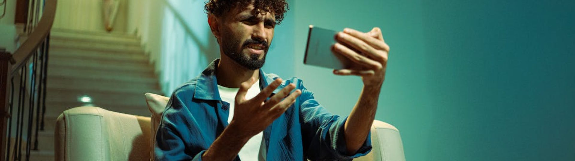 A man sitting on a couch indoors, looking confused while using a smartphone.