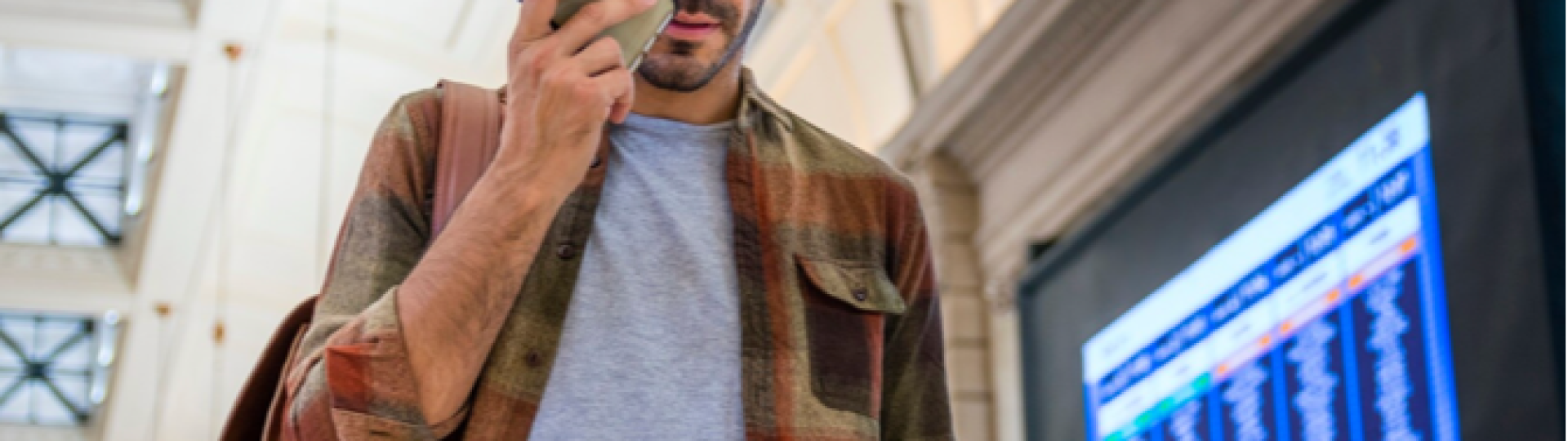 Man talking on a smartphone while reading a document near a digital travel information board