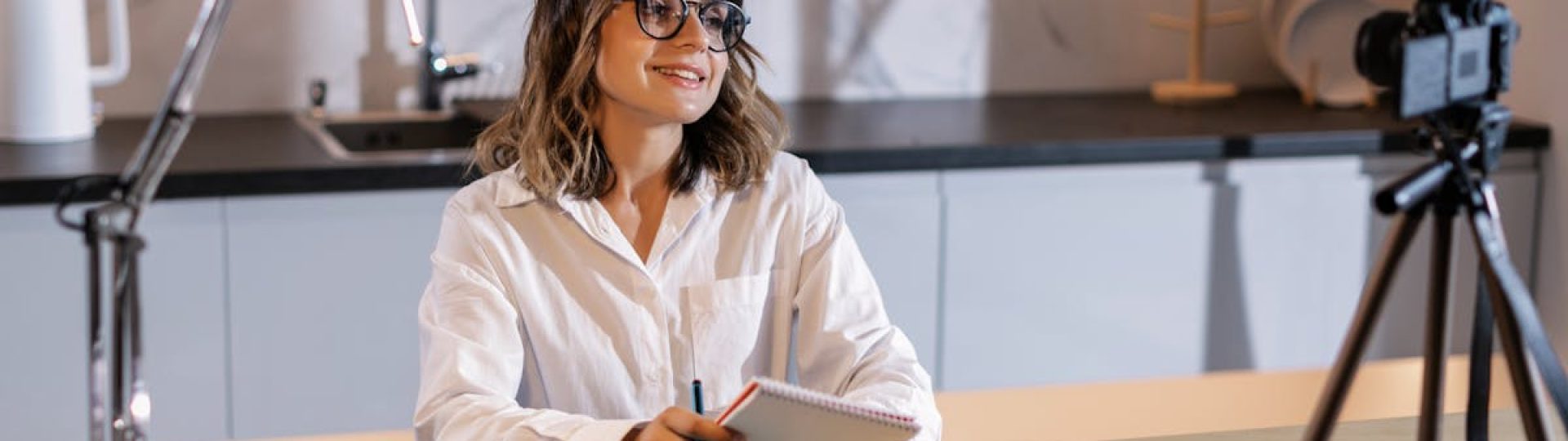 Young woman in glasses working indoors with laptop and camera, smiling while taking notes.