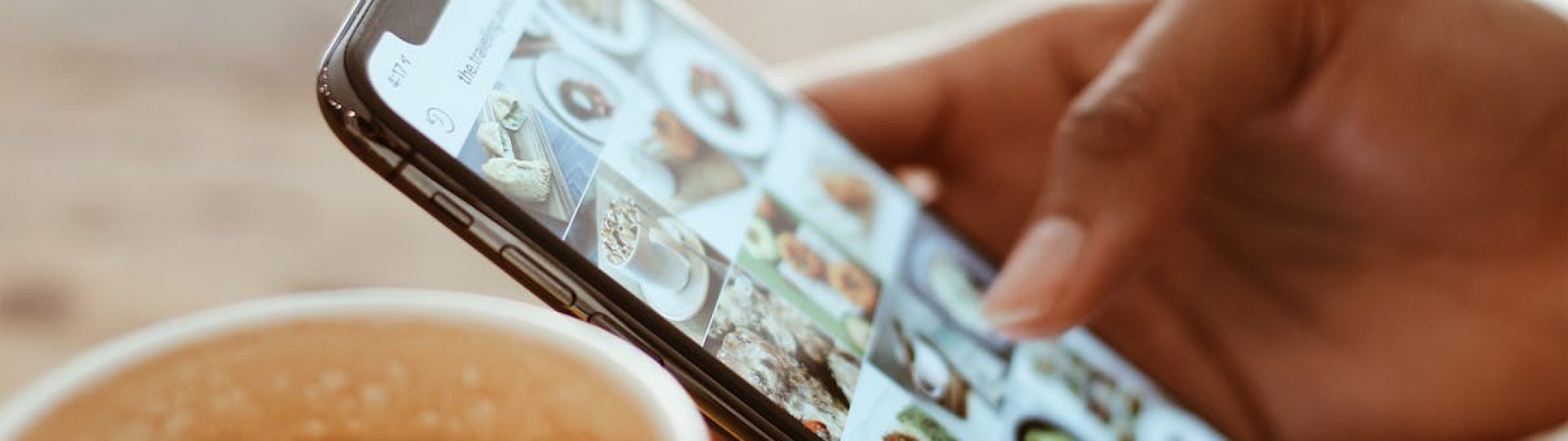Hand browsing social media photos on a smartphone next to a cup of coffee indoors.