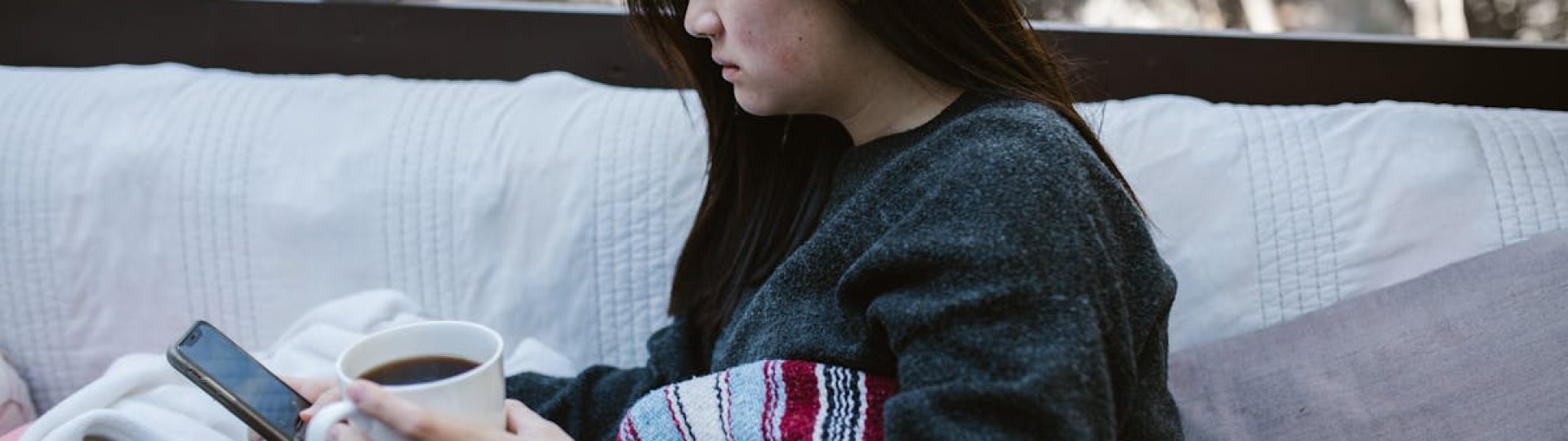 A woman enjoys a quiet moment with coffee and a phone on a cozy outdoor couch.