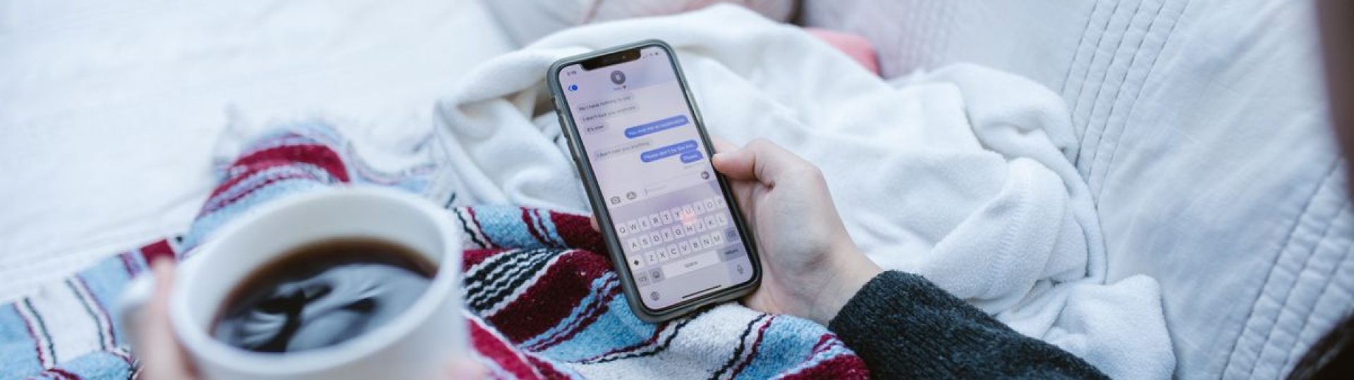 Person relaxing indoors with coffee and smartphone, texting in a cozy setting.