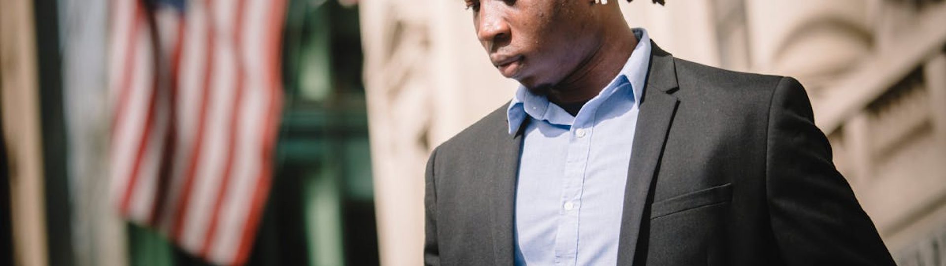 Low angle of young African American male entrepreneur in formal suit messaging on cellphone with wireless earbuds in ears while standing against blurred facade of building with American flags