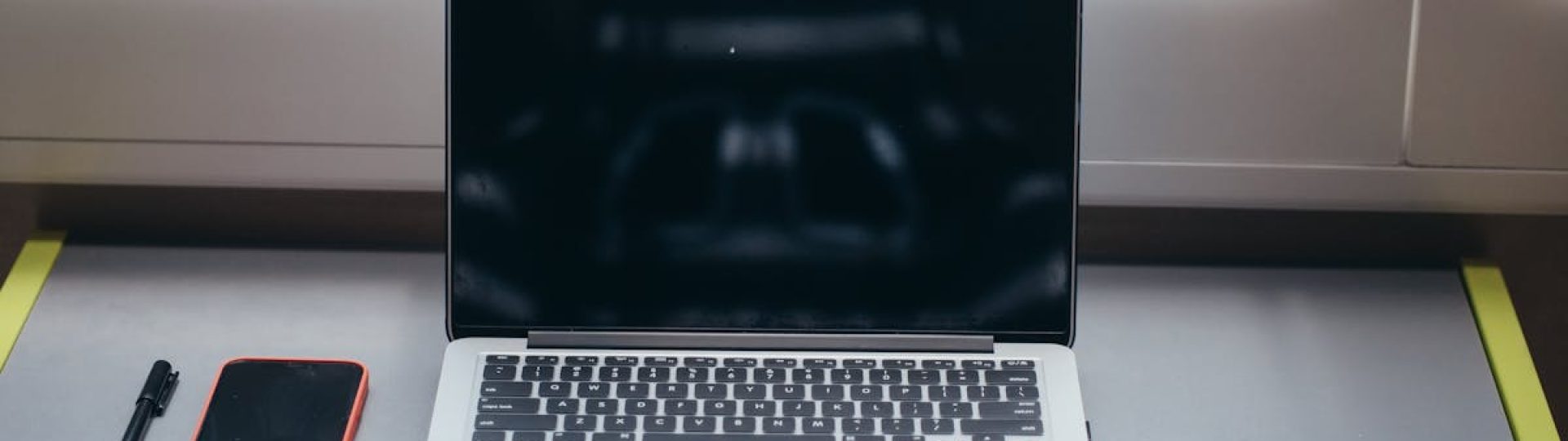 A minimalist desk setup featuring a laptop, smartphone, and pen under soft window light.