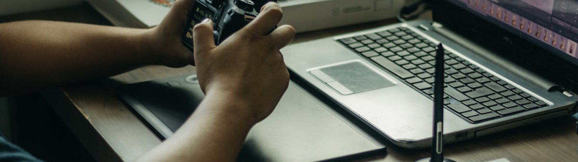 Photographer using a laptop and stylus for photo editing, holding a camera in an indoor setup.