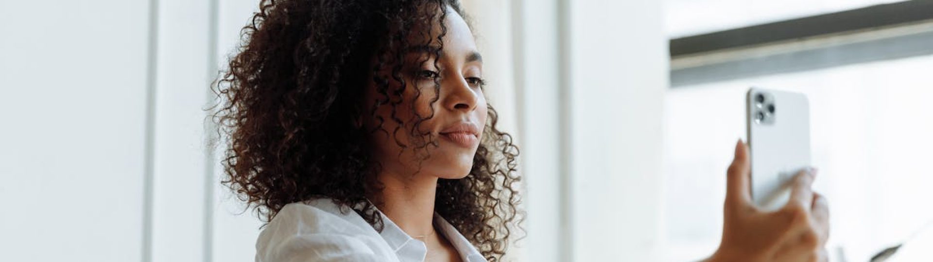 Young woman with curly hair taking a selfie with smartphone indoors.