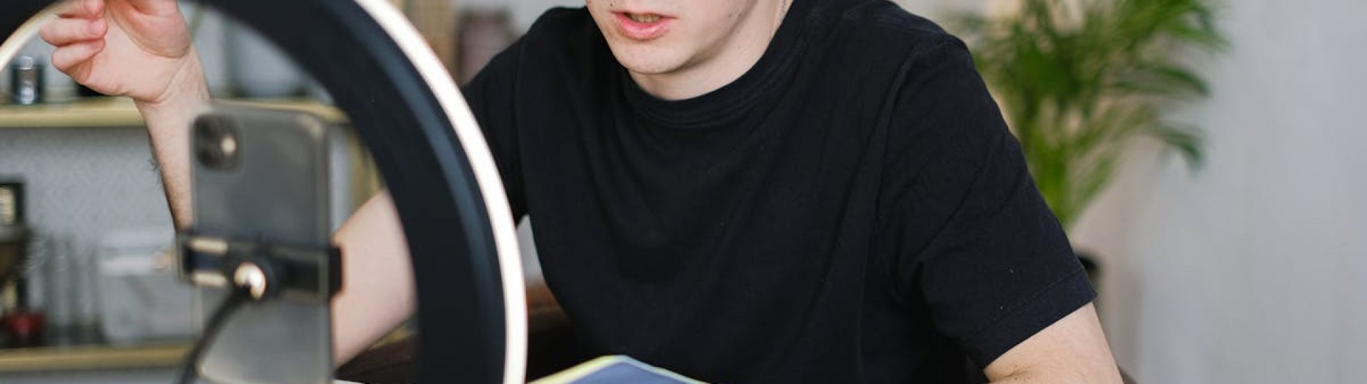 Adult male recording video content indoors, using a smartphone and ring light, holding a notebook.