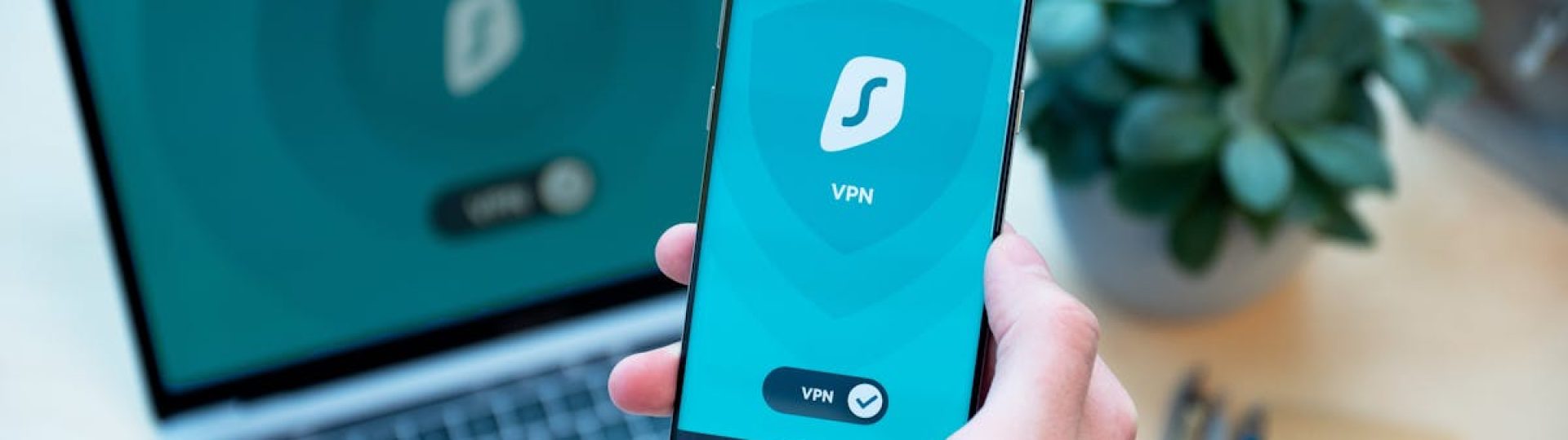 Close-up of a hand holding a smartphone with VPN app, laptop in the background, showcasing digital security.